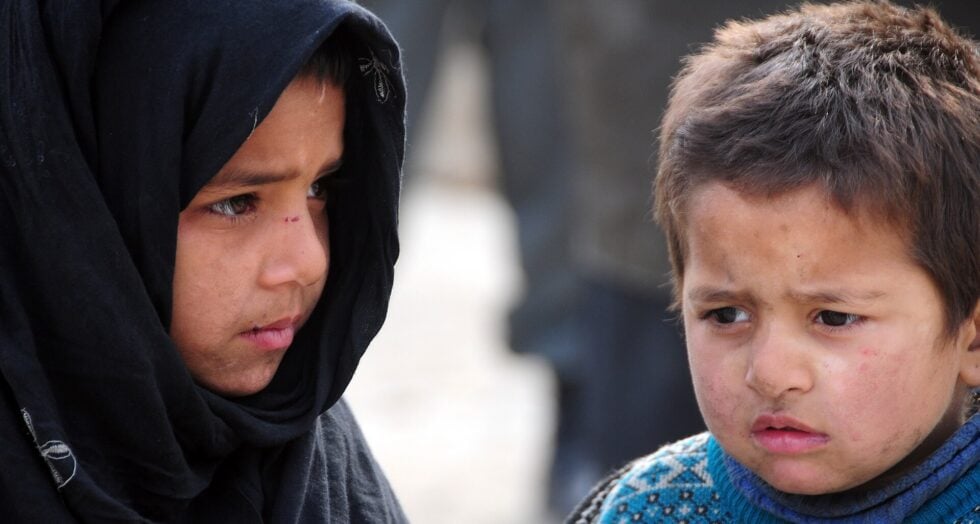 Two children stand side by side, the girl in a black scarf and the boy in a blue sweater, both looking somber.