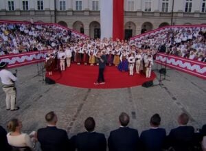 Crowd surrounding a circular red-carpet stage with people in traditional costumes and a speaker performing center stage at an outdoor ceremony.