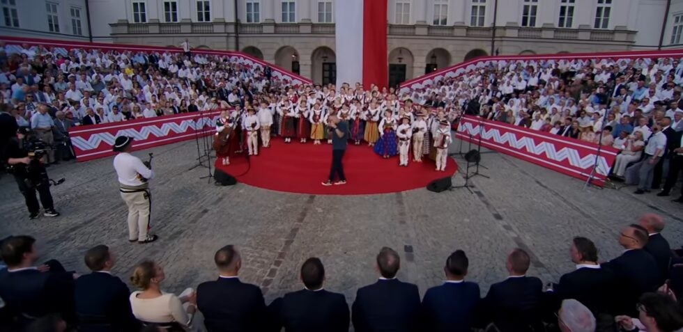 Crowd surrounding a circular red-carpet stage with people in traditional costumes and a speaker performing center stage at an outdoor ceremony.