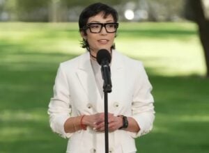 Woman in a white blazer speaks into a microphone in a sunlit park with green grass and trees behind her.