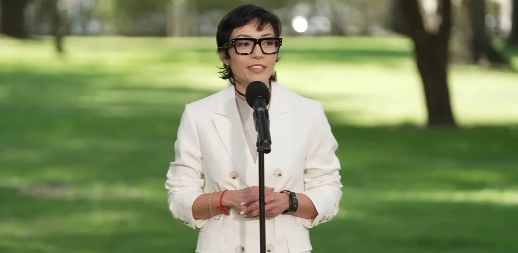 Woman in a white blazer speaks into a microphone in a sunlit park with green grass and trees behind her.