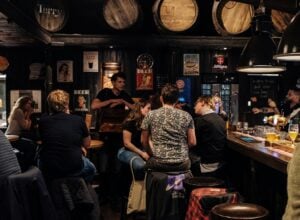 Busy rustic pub interior with customers seated at the bar and a bartender serving behind the counter, warm dim lighting, beer taps in view.