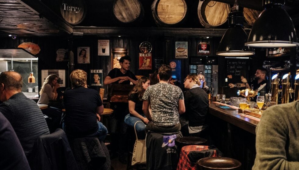 Busy rustic pub interior with customers seated at the bar and a bartender serving behind the counter, warm dim lighting, beer taps in view.