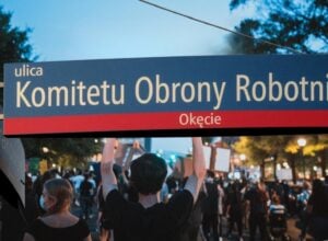 Niebieska tablica z nazwą ul. Komitetu Obrony Robotników, w tle widziany od tyłu tłum idący w proteście