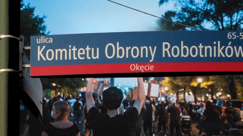 Niebieska tablica z nazwą ul. Komitetu Obrony Robotników, w tle widziany od tyłu tłum idący w proteście