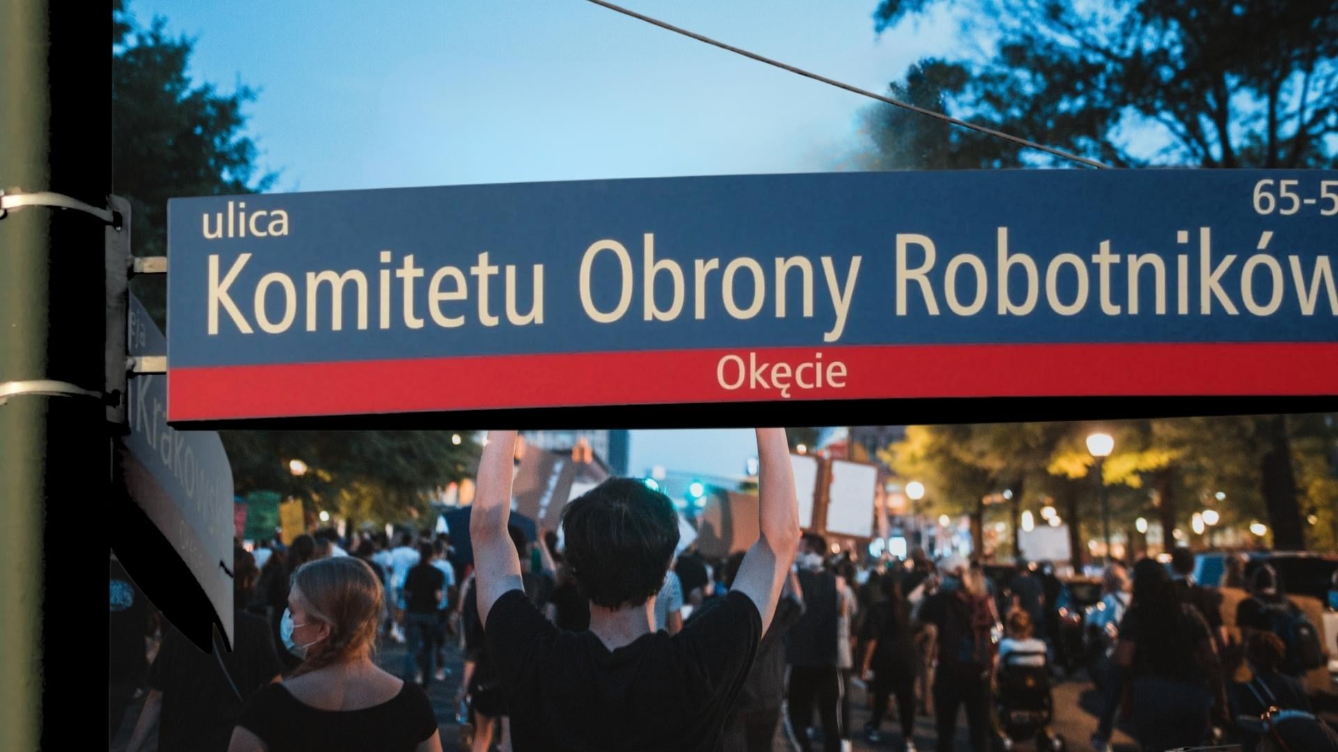 Niebieska tablica z nazwą ul. Komitetu Obrony Robotników, w tle widziany od tyłu tłum idący w proteście