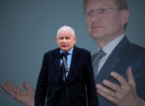 Szaro-niebieskie tło, na środku mała postać Jarosława Kaczyńskiego przy mikrofonie, za nim ogromny Leszek Balcerowicz