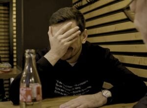 Mężczyzna w czarnej bluzie (Ilja Jaszyn) siedzi przy stoliku w knajpie i zasłania twarz prawą dłonią