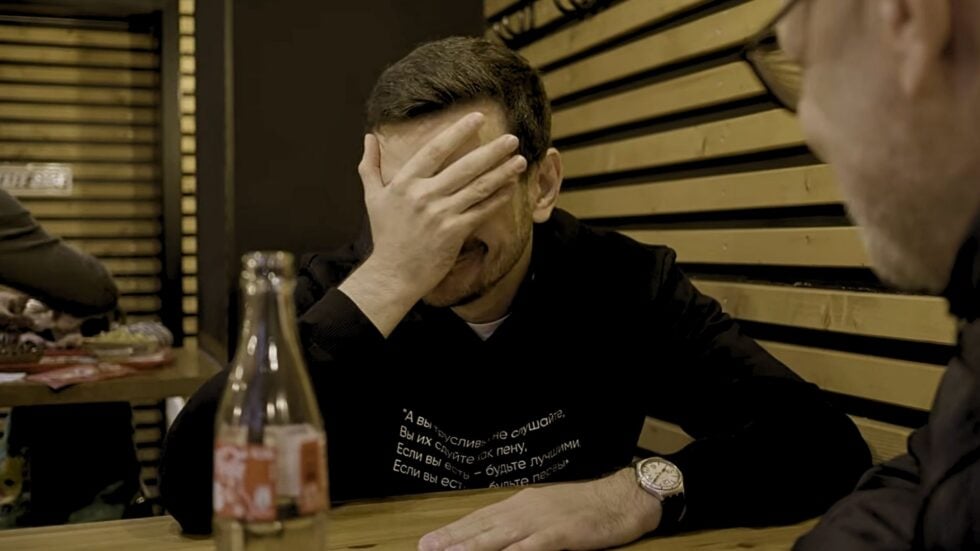 Mężczyzna w czarnej bluzie (Ilja Jaszyn) siedzi przy stoliku w knajpie i zasłania twarz prawą dłonią