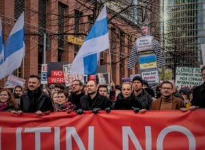 Maszerujący tłum z wielkim czerwonym banerem na przodzie, powiewają flagi rosyjskiej opozycji antywojennej (biało-niebiesko-białe), na przodzie idzie Julia Nawalna