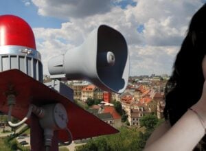 W tle lubelska starówka, na pierwszym planie po lewej głośniki z syreną alarmową, po prawej przycięta do połowy twarz przestraszonej kobiety załaniającej dłonią usta