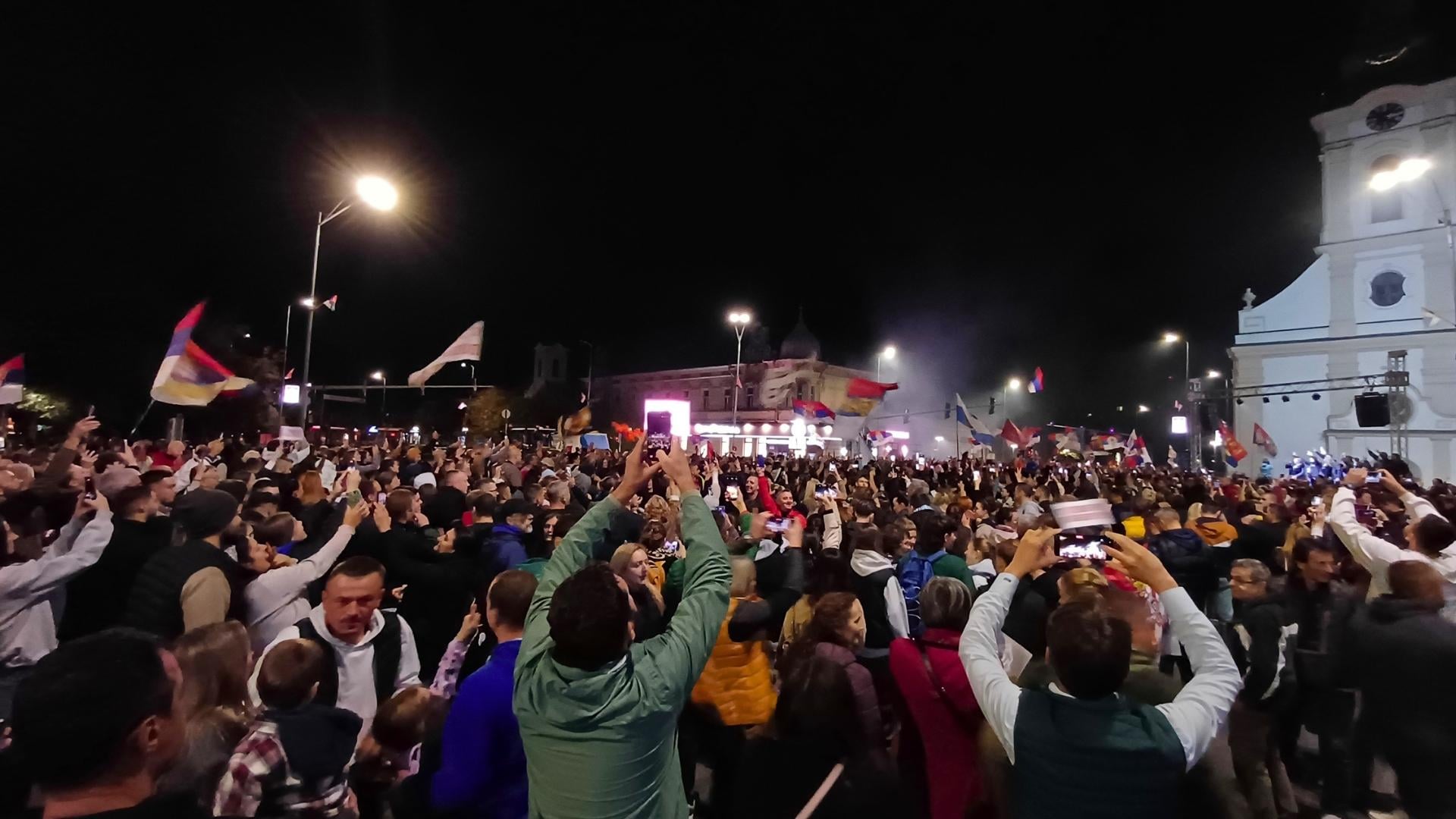Tłum protestujący nocą, nad nim widać wiele powiewających flag Serbii