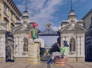 Brama Główna do kampusu centralnego Uniwersytetu Warszawskiego, przy wejściu siedzą dwie rysunkowe postaci - jedna na wysokiej kupce monet, druga na niskiej
